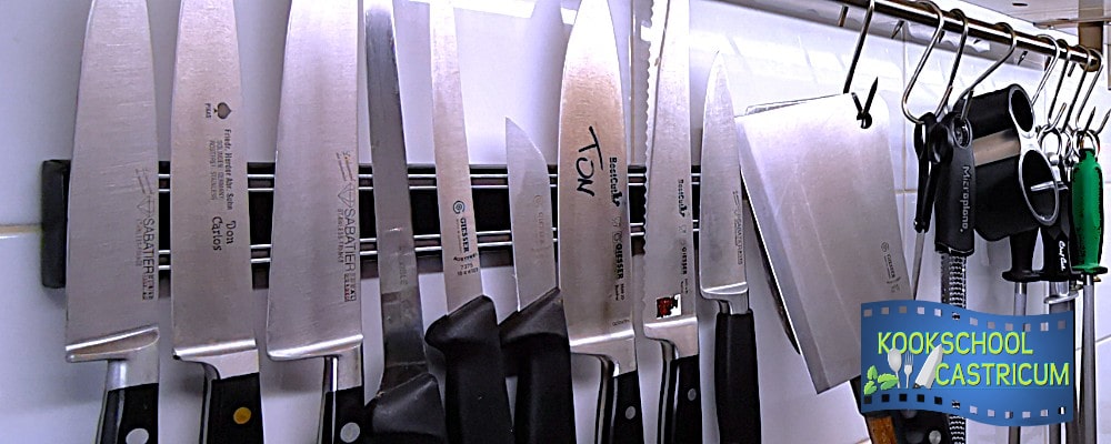Row of stainless steel kitchen knives hanging on a magnetic strip, with black handles and a blue KOOKSCHOOL CASTRICUM logo in the corner.